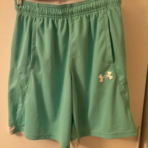 Turkosgröna träningsshorts från Under Armour - Snygga turkosgröna träningsshorts från Under Armour med vitt logotryck på benet och vita detaljer vid benslut. Shortsen har resår i midjan och är tillverkade i 100% polyester, perfekt för sport och aktivitet. Liten fläck på framsidan