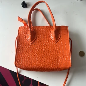 Orange axelväska med krokodilpräglat mönster - Snygg orange axelväska med krokodilpräglat mönster. Väskan har både handtag och lång axelrem samt dragkedja upptill. Perfekt för att addera färg till din outfit.