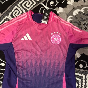 Tysklands fotbollströja i rosa och lila från Adidas - Säljer en officiell Tyskland fotbollströja från Adidas i rosa och lila med vita detaljer. Tröjan har korta ärmar, rund hals och ett grafiskt mönster framtill. Tysklands landslagsmärke och fyra stjärnor på bröstet. Perfekt för fotbollsfans!