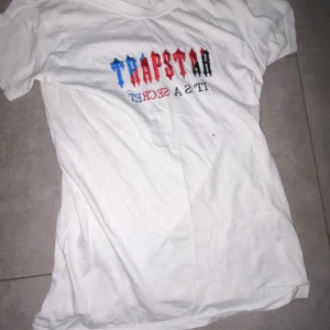 Vit t-shirt från Trapstar - Vit t-shirt från Trapstar med tryck i blått, rött och svart på bröstet. Klassisk passform och rund halsringning. Perfekt för en avslappnad streetwear-look.