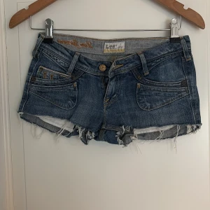 Blå jeansshorts från Lee Girl - Säljer ett par klassiska blå jeansshorts från Lee med råa, fransiga kanter och fem fickor. Shortsen har låg midja och slitna detaljer, längd fram 15cm. Barn strl 10Y