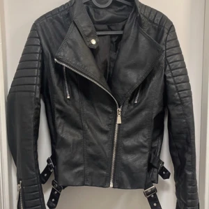 Svart skinnjacka med dragkedjor och bältesdetaljer - Säljer en svart skinnjacka med coola detaljer som snedställd dragkedja, quiltade partier på axlar och ärmar samt bälten i midjan. Jackan har flera dragkedjefickor och en klassisk bikerstil. Perfekt för dig som vill ha en edgy look.