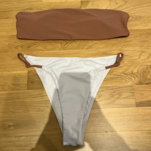 Brun och vit bikini med bandeau-topp - Säljer en snygg bikini med bandeau-topp i brun färg och högt skurna vita bikinitrosor med bruna detaljer och ringar i sidorna som man kan justera. Perfekt för stranden eller poolen!