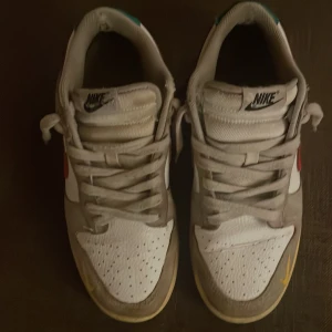 Nike Dunk Low med röd swoosh och blå sula - Nike Dunk Low sneakers med vit ovandel, gråa paneler och röd swoosh på sidorna. Skorna har blå yttersula, gula detaljer vid tån och grå snörning. Klassisk rund tå och platt sula. Jag har inte lådan till dessa! Fint skick! Priset är hugget i sten! 