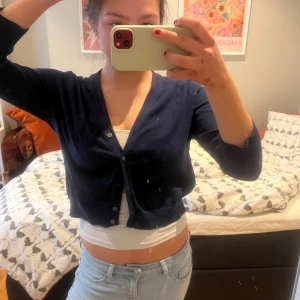 Mörkblå croppad kofta - Säljer en mörkblå croppad kofta med v-ringning och knappar framtill. Tröjan har lång ärm och är perfekt att styla över ett linne eller t-shirt. Enkel och stilren design som passar till mycket.