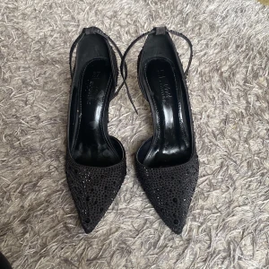 Svarta glittriga pumps med spetsig tå - Säljer ett par svarta pumps med spetsig tå och klack. Skorna är täckta av svarta glittriga paljetter och har en smal rem runt vristen. Helt nya, aldrig använda💖