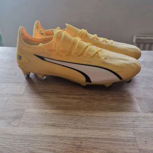 Gula Puma Ultra fotbollsskor - Säljer ett par gula Puma Ultra fotbollsskor med svarta och vita detaljer. Skorna har snörning, låg profil och mönstrad sula för bra grepp på planen. Lätta och smidiga med svensk flagga på hälen och texten 'ERABI' på sidan. Alltså Hammarby if’s anfallare jusef erabi.