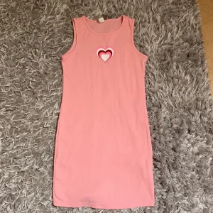 Rosa ärmlös ribbad klänning med hjärtdetalj - Säljer en söt rosa ärmlös klänning med ribbad struktur och ett tryckt hjärta i vitt och rött framtill. Klänningen är figurnära och har en enkel, rak passform som passar perfekt till sommaren.