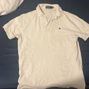 Vit pikétröja från Polo Ralph Lauren - Klassisk vit pikétröja från Polo Ralph Lauren med liten broderad logga på bröstet. Tröjan har krage och knappar vid halsen, perfekt för en clean och stilren look.