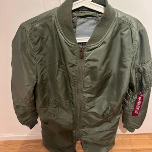 Grön bomberjacka från Alpha Industries - Säljer en klassisk grön bomberjacka från Alpha Industries med dragkedja framtill, ribbade muddar och fickor med tryckknappar. Jackan har en ikonisk röd tag på ärmen och en ärmficka med dragkedja.