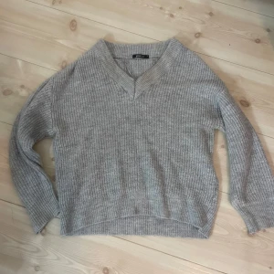 Grå stickad tröja med v-ringning från Gina  - Mysig grå stickad tröja med v-ringad hals och avslappnad passform. Tröjan har långa ärmar och ribbad struktur, perfekt för kyligare dagar. 
