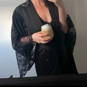 Svart kimono från Hunkemöller - Elegant svart kimono från Hunkemöller med vackra spetsdetaljer på ärmarna och axlarna. Kimonon har ett knytband i midjan och är tillverkad i ett mjukt, glansigt material som ger en lyxig känsla.