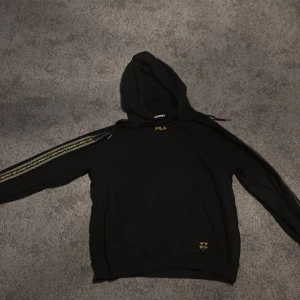 Svart hoodie från Fila med gulddetaljer - Svart hoodie från Fila med guldfärgad logga på bröstet och dekorativa gulddetaljer längs ena ärmen. Klassisk huva och avslappnad passform, perfekt för en sportig och casual stil.
