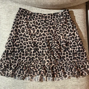 Leopardmönstrad kjol med volang från Lindex - Säljer en kort leopardmönstrad kjol från Lindex med volangdetaljer längs nederkanten och omlottkänsla framtill. Kjolen har resår i midjan och är i ett lätt, luftigt material. Perfekt för dig som gillar djurmönster och vill sticka ut.