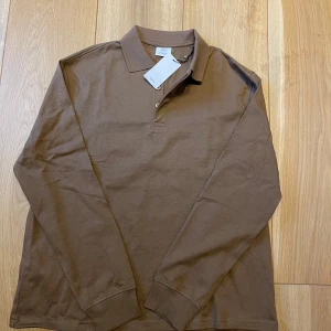 Brun långärmad pikétröja från mango - Säljer en stilren brun långärmad pikétröja från Arket. Tröjan har klassisk krage och knappar vid halsen. Perfekt för en avslappnad men ändå uppklädd look.