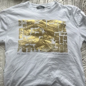 Vit t-shirt med guldtryck från Zara - Vit t-shirt från Zara med ett stort abstrakt guldmönster på framsidan. T-shirten har rund halsringning och korta ärmar. Storlek S, passar även M då den är lite oversized! Använd max 2 gånger