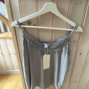 Grå linnetopp med spetsdetaljer  - Säljer en stilren grå linnetopp med tunna axelband och fina spetsdetaljer längs urringningen. Perfekt att matcha med jeans eller kjol för en trendig look.
