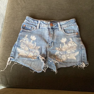 Blå jeansshorts med broderade blommor - Säljer ett par blå jeansshorts från Lindex med snygga fransar och vita broderade blommor framtill. Shortsen har hög midja, fem fickor och råa kanter för en cool look. Perfekta för sommaren!