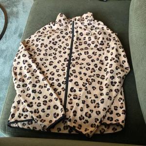 Leopardmönstrad fleecejacka från H&M - Säljer en beige fleecejacka från H&M med leopardmönster och hel dragkedja framtill. Jackan har lång ärm och hög krage, perfekt för att hålla värmen under kyliga dagar. Mjuk och skön att ha på sig.