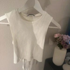 Vit ribbad croppad linnetopp med knyt - Trendig vit ribbad linnetopp med croppad passform och öppen rygg med knytning. Toppen är ärmlös och har en öppen detalj i ryggen som ger en cool look. Perfekt för varma dagar eller lager på lager-stil.