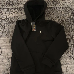 Svart hoodie från Polo Ralph Lauren - Säljer en svart hoodie från Polo Ralph Lauren med klassisk broderad logga på bröstet. Tröjan har huva med dragsko och en stor magficka framtill. Perfekt för en avslappnad och stilren look.