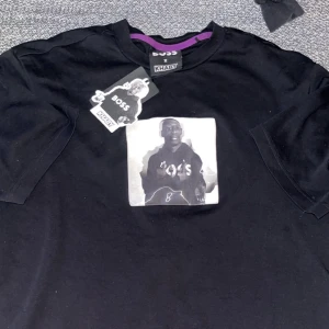 Svart t-shirt BOSS x Khaby - Svart t-shirt från BOSS x Khaby med tryck av Khaby på bröstet. T-shirten har rund hals och korta ärmar. Perfekt för dig som gillar streetwear och samarbeten.