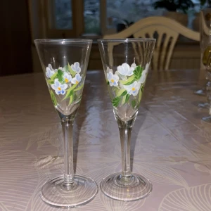 Handmålade snapsglas med blommor - Två smala snapsglas i klarglas med handmålat blommönster i vitt, blått och grönt. Glasen har en klassisk form med hög fot och konisk kupa, perfekt för snaps eller likör.