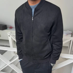 Svart bomberjacka med dragkedja - Säljer en stilren svart bomberjacka med dragkedja framtill. Jackan har ribbade muddar vid ärmslut, krage och nederkant. Passformen är normal och materialet känns mjukt och smidigt. Perfekt för dig som vill ha en klassisk look.