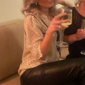 Prickig blus med svarta detaljer - Snygg vit blus med svarta prickar och svarta detaljer längs kanterna. Blusen har knappar framtill och en dekorativ rosett vid halsen. Perfekt att styla med skinnbyxor för en trendig look.