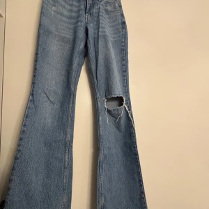 Blå bootcut jeans med hål på knät - Säljer ett par blå bootcut jeans med ett stort hål på höger knä. Jeansen har klassisk femficksdesign och är tillverkade i denim. Använda ett få par gånger💞