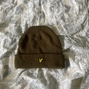 lyle&scott mössa - Säljer en olivgrön stickad mössa från Lyle & Scott med gul broderad logga framtill. Klassisk modell med uppvikt kant, perfekt för kyliga dagar.