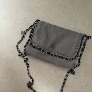Grå axelväska från Stella McCartney - Säljer en snygg grå axelväska från Stella McCartney med kedjedetaljer runt kanterna och på axelremmen. Väskan har lockstängning med magnet och ett innerfack med dragkedja. Perfekt för dig som gillar stilrena accessoarer med en cool touch.