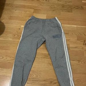 Grå mjukisbyxor från Nike med vita ränder - Grå mjukisbyxor från Nike med vita ränder längs sidorna och Nike-logga på vänster lår. Byxorna har resår i midjan och är perfekta för en avslappnad stil.