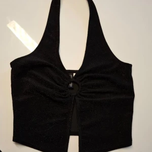 Svart glittrig halterneck topp - Säljer en svart glittrig halterneck topp från H&M Divided i storlek XS. Toppen har en snygg ringdetalj framtill med rynk och en liten slits under. Perfekt för att sticka ut med en trendig look.