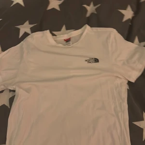 Vit t-shirt från The North Face - Säljer en klassisk vit t-shirt från The North Face med liten svart logga på bröstet. T-shirten har rund hals och korta ärmar, perfekt för en clean och enkel stil. Passar till det mesta och är riktigt skön att bära.