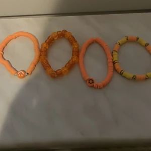 Fyra färgglada armband med pärlor och smileys - Fyra elastiska armband i orange och gult med olika pärlor och smiley-detaljer. Perfekta för att addera färg till din stil. Armbanden har runda och platta pärlor i plast och passar de flesta handleder.