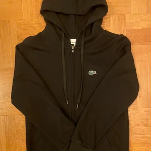 Lacoste zip up Svart  - Säljer en svart hoodie från Lacoste med dragkedja och huva. Perfekt för en chill och stilren look. Den är bara andvänd 1 gång så typ ny skick. Priset går att diskuteras vid snabb äffar. Vid frågor är det bara att skriva:)