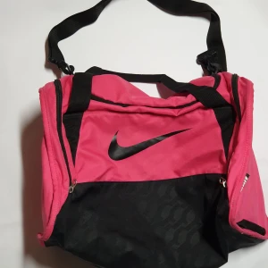 Rosa och svart träningsväska från Nike - Säljer en sportig rosa och svart träningsväska från Nike med justerbar axelrem och dragkedja. Perfekt för gymmet eller sportaktiviteter. Snygg design med stor Nike-logga på framsidan.