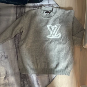 Grå stickad tröja från Louis Vuitton - Säljer en grå stickad tröja från Louis Vuitton med stor vit LV-logga på bröstet. Tröjan har rund halsringning och ribbade muddar vid ärmslut och nederkant. Perfekt för dig som gillar exklusiva märken.
