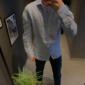 Ljusblå randig skjorta från Bläck - Snygg ljusblå skjorta från Bläck med vita ränder och klassisk krage. Skjortan har lång ärm, knappar framtill och är i en skräddad passform (tailored fit). Perfekt för dig som gillar stilrena och fräscha plagg.