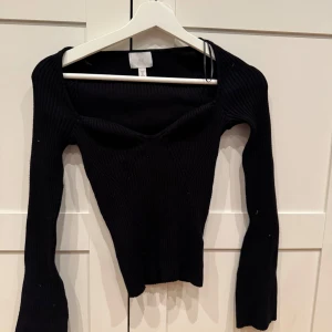 Svart ribbad topp från H&M - Säljer en svart ribbad topp från H&M med lång ärm och djup v-ringning. Toppen är tight och har en snygg, figurnära passform som framhäver siluetten. Perfekt att styla med jeans eller kjol för en trendig look.