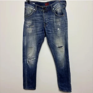 Blå jeans med slitningar  - Säljer ett par blå jeans med slitningar och hål på framsidan. Jeansen har fem fickor, normal passform och raka ben. De har en klassisk knappgylf och är tillverkade i denim. Perfekta för en avslappnad stil. ‼️Öppen för byten och även sänka priset‼️