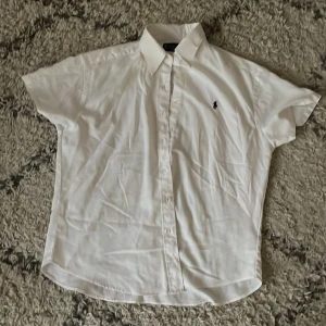 Vit kortärmad skjorta från Ralph Lauren - Säljer en klassisk vit kortärmad skjorta från Ralph Lauren med broderad logga på bröstet. Skjortan har knappar framtill och är tillverkad i 100% bomull. Perfekt för en stilren och avslappnad look. Vänligen ställ några frågor om du undrar något.