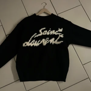 Svart stickad tröja från Saint Laurent - Säljer en svart stickad tröja från Saint Laurent med vit, stor logotyp på bröstet. Tröjan har lång ärm och en klassisk, avslappnad passform. Perfekt för dig som gillar exklusiva och trendiga plagg. Pris kan diskuteras, vid frågor tveka inte på att höra av dig