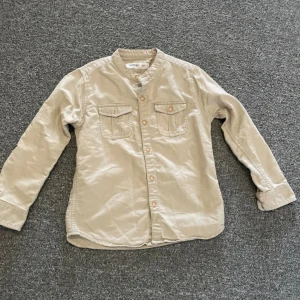 Beige skjorta med ståkrage från Redtag - Snygg beige skjorta från Redtag med ståkrage och två bröstfickor med lock. Skjortan har långa ärmar och knäppning framtill. Perfekt för en stilren look.