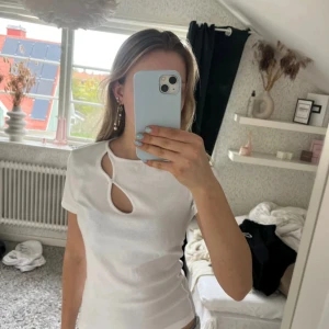 Vit ribbad topp med cut-out detalj - Säljer en vit ribbad topp med kort ärm och unik cut-out detalj framtill. Toppen har en liten knapp vid öppningen och är figurnära i modellen. Perfekt för dig som vill ha något stilrent men ändå med en twist.