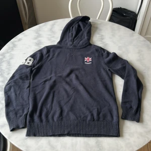 Mörkblå stickad hoodie från Morris - Säljer en mörkblå stickad hoodie från Morris med broderad logga och siffror på ärmen. Tröjan har långa ärmar, ribbade muddar och huva. Perfekt för kyliga dagar och snygg till jeans. Bra använt skick. 