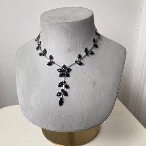 Vintage halsband med svarta stenar - Säljer ett elegant halsband med blommor av svarta, facetterade stenar och små silverfärgade detaljer. Halsbandet har en justerbar kedja baktill och en unik design där blommorna hänger i en v-formad rad framtill.