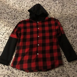 Röd och svart rutig skjorta med huva från SHEIN - Säljer en röd och svart rutig skjorta från SHEIN med svarta ärmar och huva. Skjortan har knappar framtill och är långärmad. Perfekt för dig som gillar en avslappnad stil med lite edge.