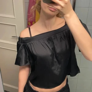 Svart offshoulder topp med volang - Säljer en svart offshoulder topp med volangdetaljer och axelband. Toppen har en lös passform och är tillverkad i ett glansigt material som ger en snygg look. Perfekt att styla med både jeans och kjol.